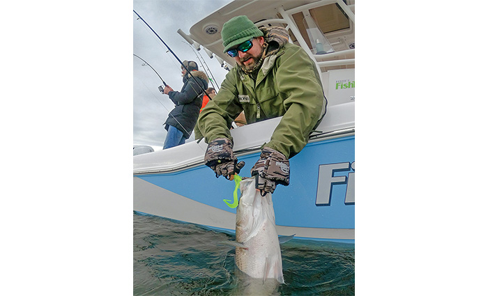 Adult male dressed in green layers pulling a a hook out of a large fish caught aboard a fishing boat.