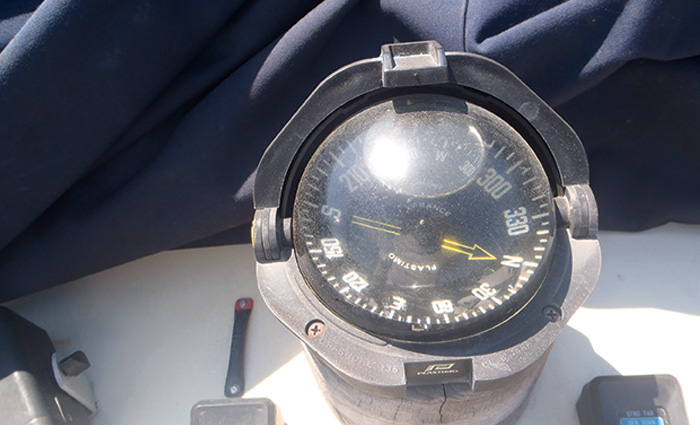 Cloudy and damaged black boat compass with white numbers and letters and a yellow arrow pointing north.