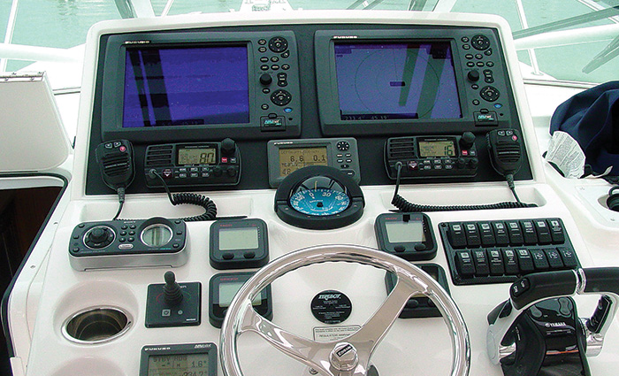 A boat compass in the center of a white navigation system with several electrical features and a silver steering wheel.