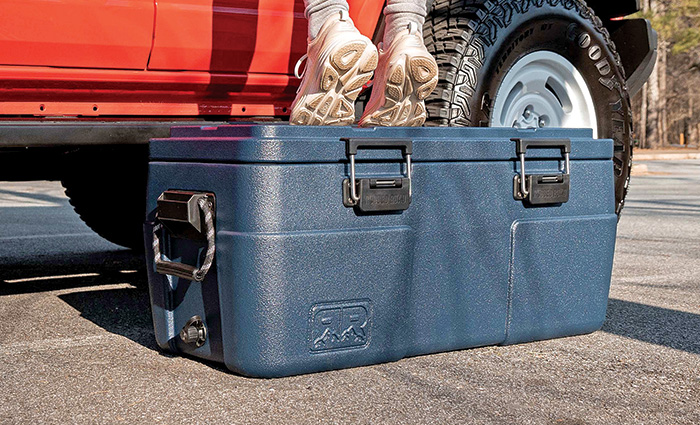 Up-close view of a person in white shoes standing on top of a large navy cooler with multiple black handles.