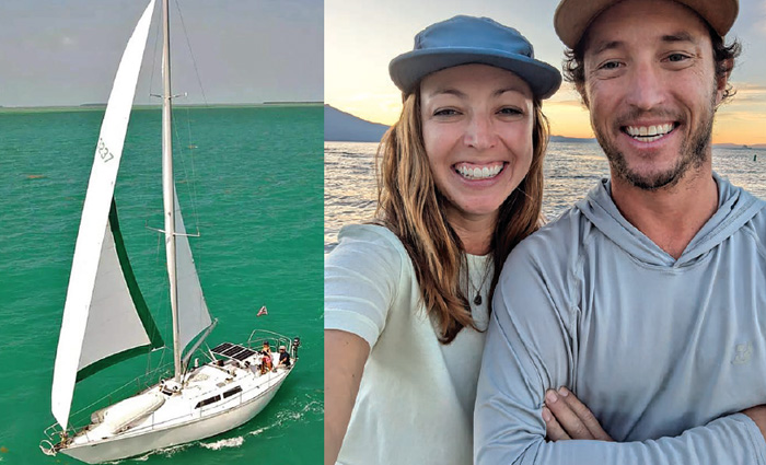 Ariel view of a white sailboat on open waters next to a Caucasian couple posing for a picture.