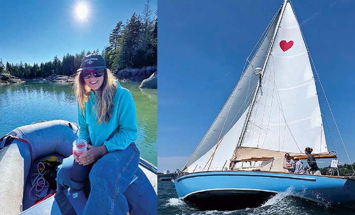 Young adult female wearing a light blue sweatshirt, jeans, sunglasses and a navy ballcap sitting on the front of a boat next to a white sailboat, with white sails and a single red heart.