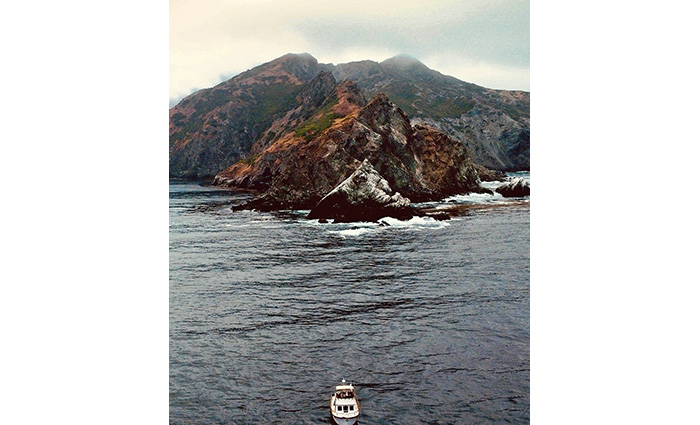 Aerial view of a white vessel departing a mountainous island with overcast skies. 