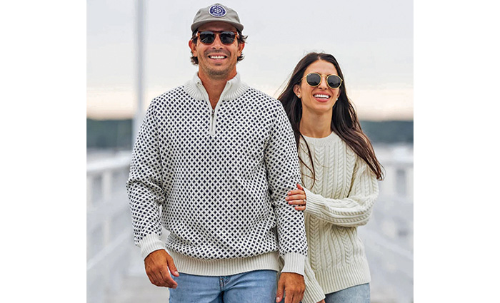An adult couple wearing sweaters and sunglasses walking along a dock at sunset.