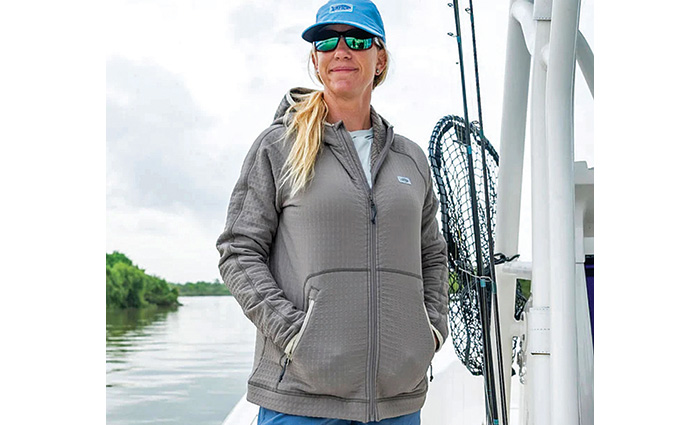 Adult female wearing a gray hooded sweatshirt, blue hat and sunglasses standing on a white vessel.