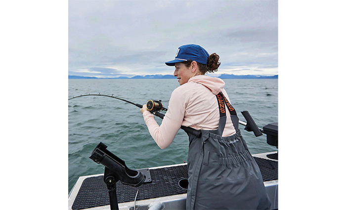 Female wearing a blue ballcap, pink hooded sweatshirt fishing aboard a silver boat on an overcast day.
