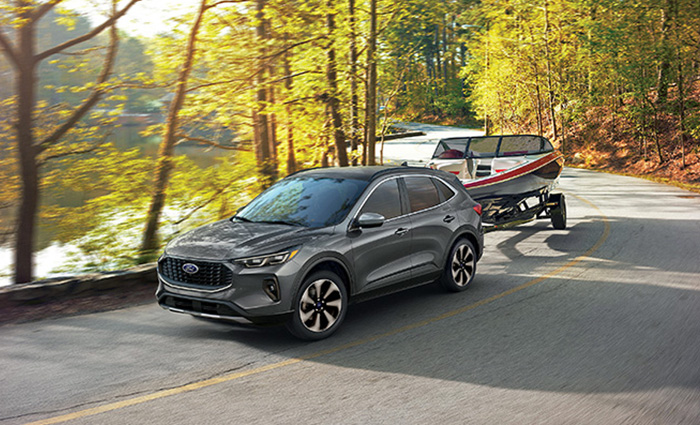 A silver SUV pulling a white and red speedboat on a road alongside a lake during fall.