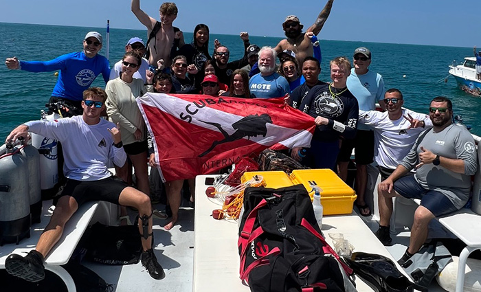 A group of young adults on a large boat on open waters during a sunny day displaying a red and white scuba flag.