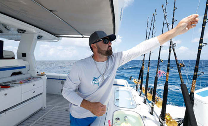 Adult male wearing a white shirt, blue shorts, ballcap and sunglasses adjusting a fishing line on a white boat.