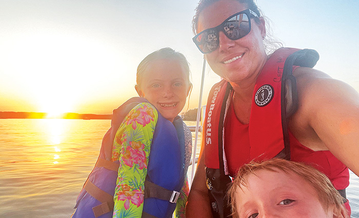 Adult female wearing black sunglasses, a red swimsuit and red life jacket taking a selfie with two kids at sunset on open waters.