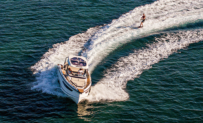 Aerial view of a white vessel speeding through blue waters while a young adult male skies behind. 