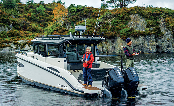 An adult male wearing a navy knit hat, plaid shirt and green life jacket and a young adult wearing a red coat and blue life jacket fishing off the back of a white boat.