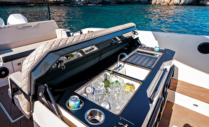 Folding bench on the back of a white boat opened and displaying a cooler filled with ice and beverages, sing and grill.