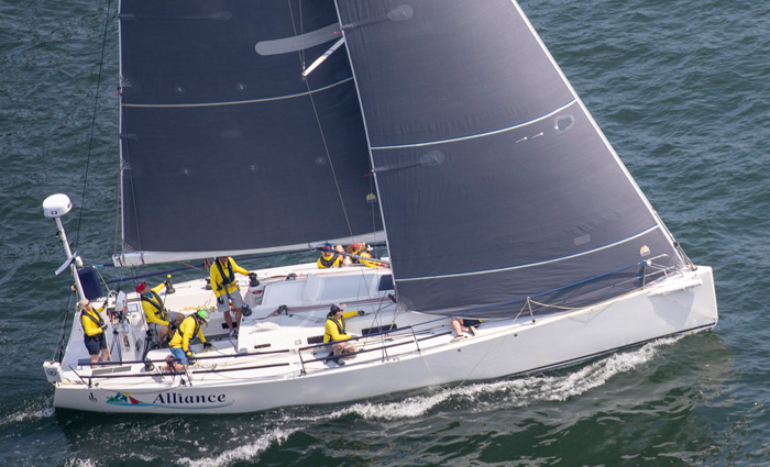 Aerial view of large white sailboat with gray sails steered by various crew members wearing yellow jackets out on open waters.