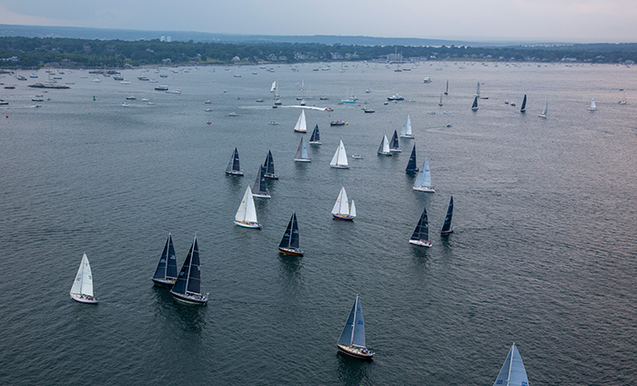 View from above of numerous white, navy blue and gray sail boats out on the water with other boats and the shoreline in the background. 