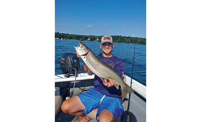 Young adult male wearing royal blue swim shorts, a navy shirt and gray hat holding a large fish on the back of a boat.