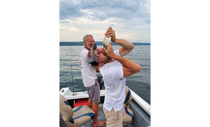 A bearded adult male wearing a white shirt and gray shorts laughing at a young adult male pretending to eat a small fish caught on the end of a boat.