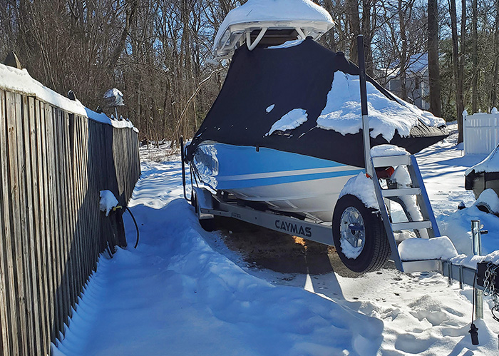 White boat on a trailer with navy cover in a snow-covered driveway.
