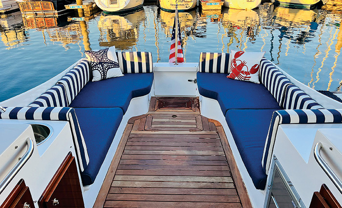 Stern of a boat on a dock with wood flooring, navy cushions, navy and white stripped pillows and an American flag.
