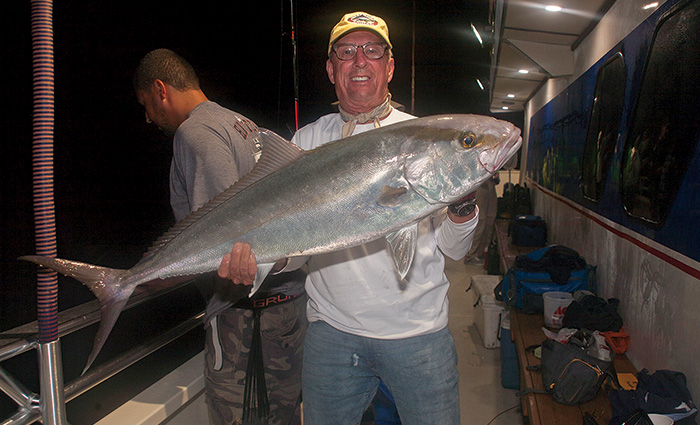 Adult male wearing glasses, white shirt, yellow hat and jeans holding a large fish on a boat at night.