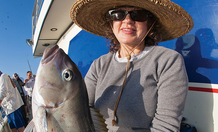 Adult female wearing sunglasses, a large straw hat and gray sweatshirt holding a big gray fish.