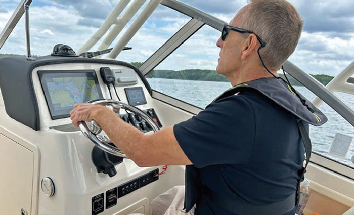 Man wearing a navy shirt and black sunglasses steering a white boat out on the water.