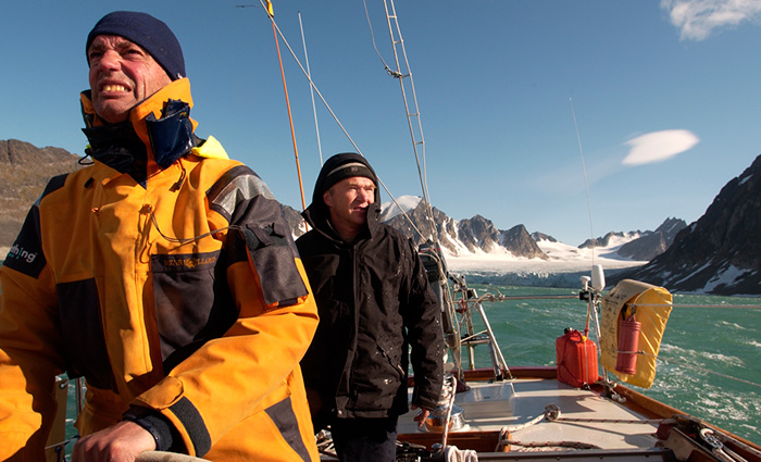 Adult male in a black stocking cap and black jacket behind a man with a navy stocking cap and yellow and black jacket steering a vessel with snow covered mountains in the background.