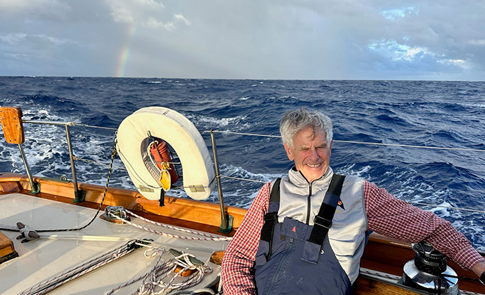 Adult male with gray hair wearing a gray vest and navy overalls sitting on the rail of a boat out at sea.