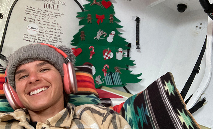 Young adult wearing a gray knit hat and pink headphones resting inside the cabin of a boat with a paper Christmas Tree on the wall.