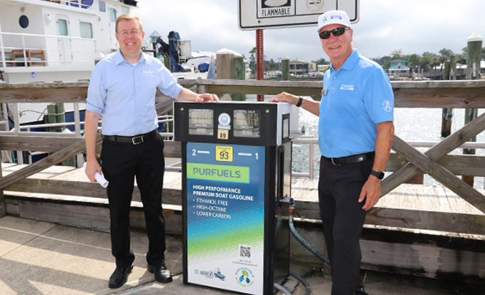 Two adult males, wearing black dress pants and blue shirts posing for a photo next to a gas tank on a wooden dock.