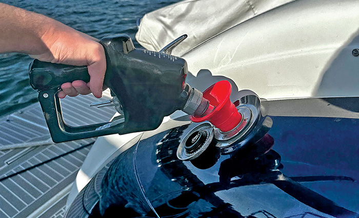 Close-up of a hand pumping gas into a navy blue boat with several vessels in the background on open waters on a clear day.
