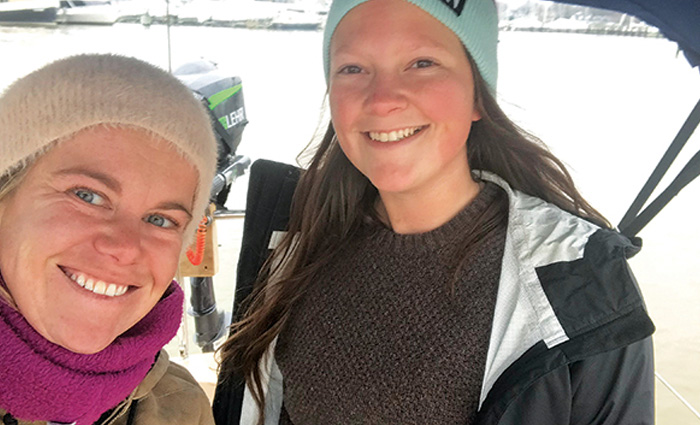 Two young adult females wearing winter clothes posing for a selfie photo while on a boat out at sea.