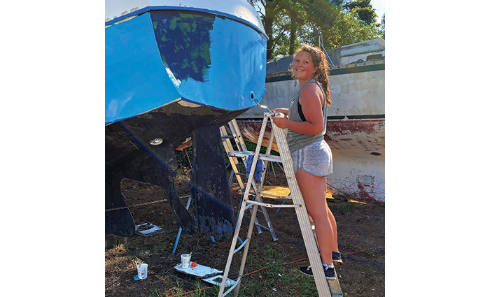 Young adult female wearing a gray tank top and gray shorts standing on a silver ladder applying blue paint to a small boat.