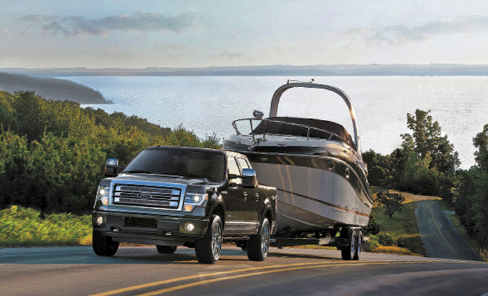 A Ford four door black truck towing a large white and black vessel up a hillside road with a big lake in the background.
