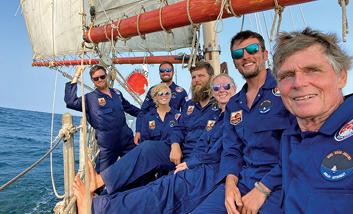 Group of adults wearing blue space jumpsuits on a large sailboat out on open waters.