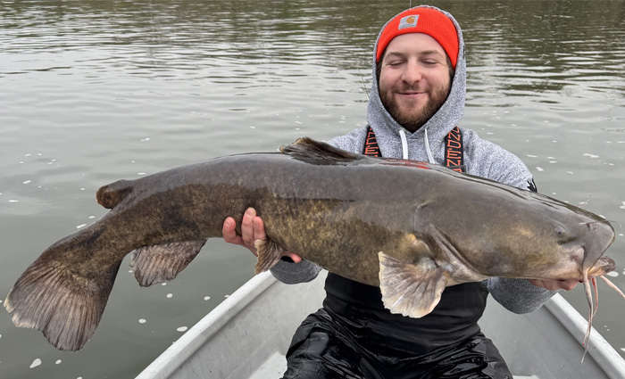 Adult male wearing an orange knit hat and gray hooded sweatshirt proudly holding a large catfish on a small fishing boat.