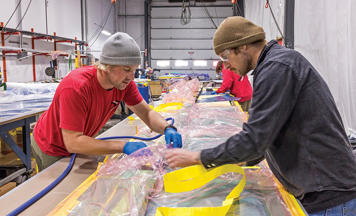 Two adult males, one wearing a gray knit hat, short sleeve red shirt and blue rubber gloves across from the other wearing a tan knit hat, safety glasses and a long sleeve black shirt working inside a warehouse.