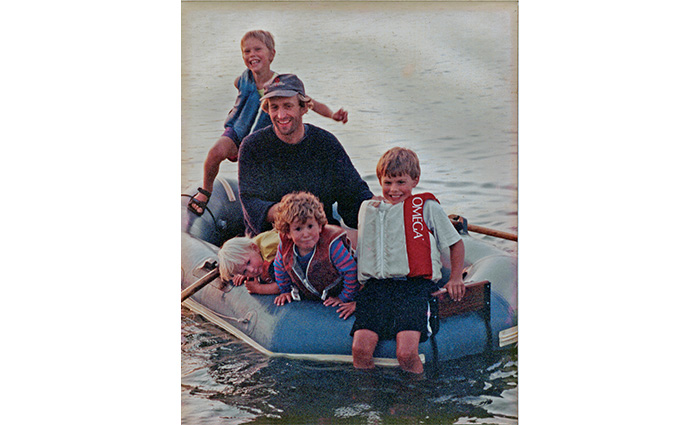 Adult male with three young boys aboard a small blue paddle boat out on the water.