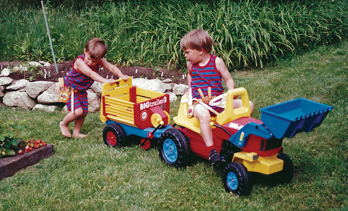 Two young boys, both wearing matching red and blue stripped outfits, playing on a toy dump truck outside on the grass.