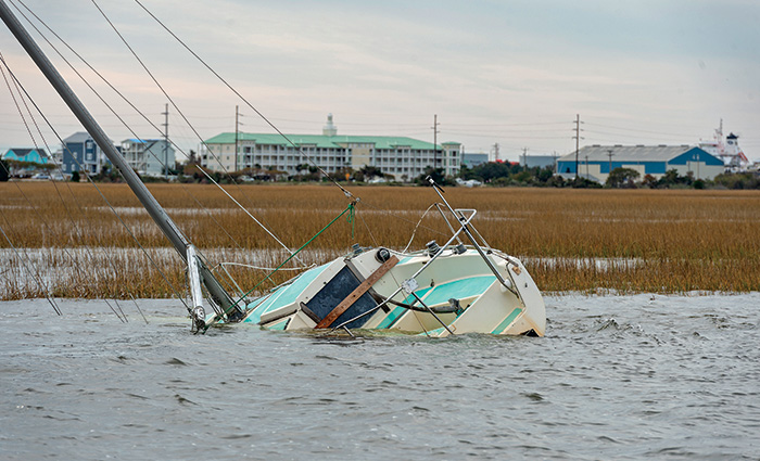A sinking white and light blue sailboat with a shoreline and buildings in the background at sunset.