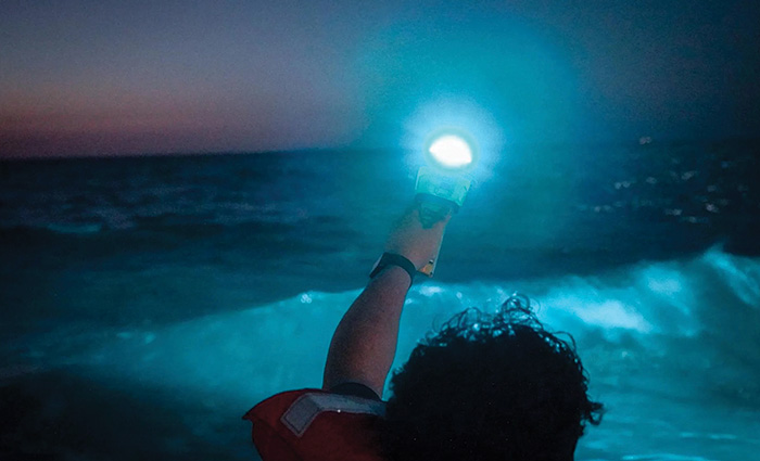 View from behind of a young adult wearing a orange life jacket holding a bright flare as a wave crashes in the background at sunset.