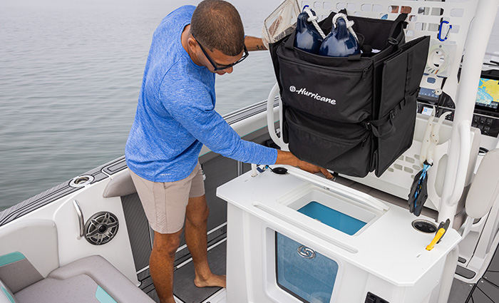 Adult male wearing a long sleeve blue shirt, black sunglasses and tan shorts holding a large black bag on a boat on the water.
