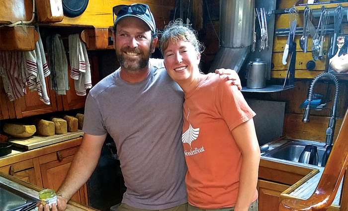 Bearded adult male wearing a gray shirt and tan shorts with his arm around an adult female wearing an orange shirt in a kitchen.