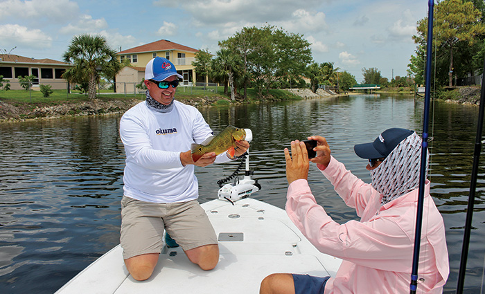 Adult male wearing a white and blue ballcap, white shirt and tan shorts showing a fish caught on the bow of a white boat while another adult male wearing a pink shirt takes a picture.