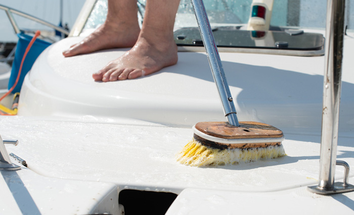 Feet of an adult next to a blue broom cleaning the top of a white boat.