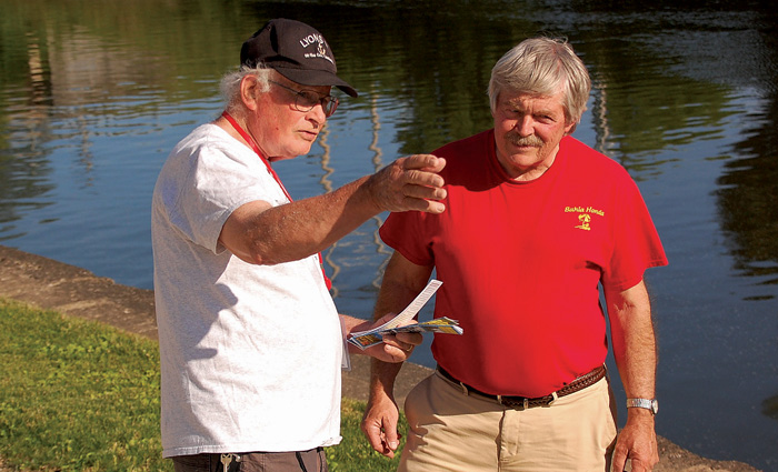 An senior male wearing a white shirt and black hat pointing out something to another senior male in a red shirt.