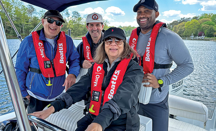 Two adult Caucasian males, an African American male, and an adult female all wearing red life jackets while posing for a photo on a boat on the water.