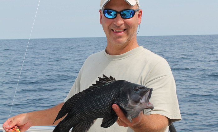 Adult male wearing a gray shirt, white hat and sunglasses proudly displaying a fish caught with open waters in the background.