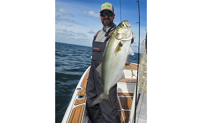 Adult bearded bale wearing a green hat, sunglasses, gray shirt and black overalls proudly displaying a large fish caught.