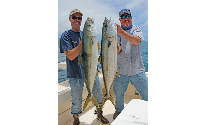 Two adult males wearing ball caps, sunglasses and jeans holding two large fish on a white boat in open waters.
